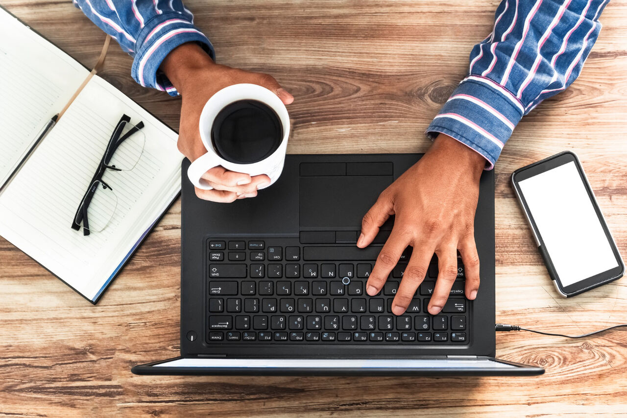 męskie dłonie na tle klawiatury laptopa, w jednej dłoni znajduje się kubek z kawą