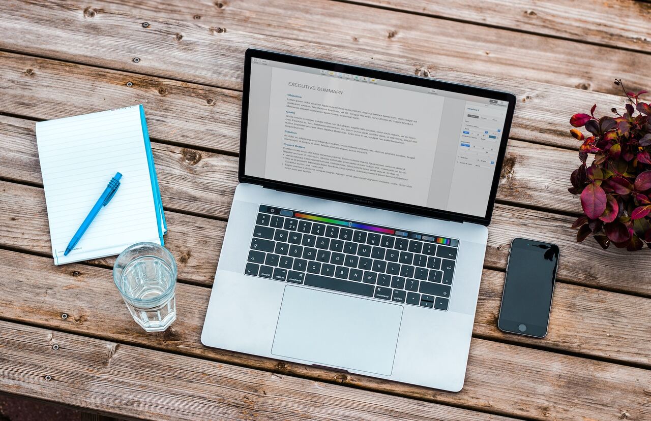 laptop leżący na stole, obok smartfon, notes i szklanka wody, proces tworzenia opisów produktów